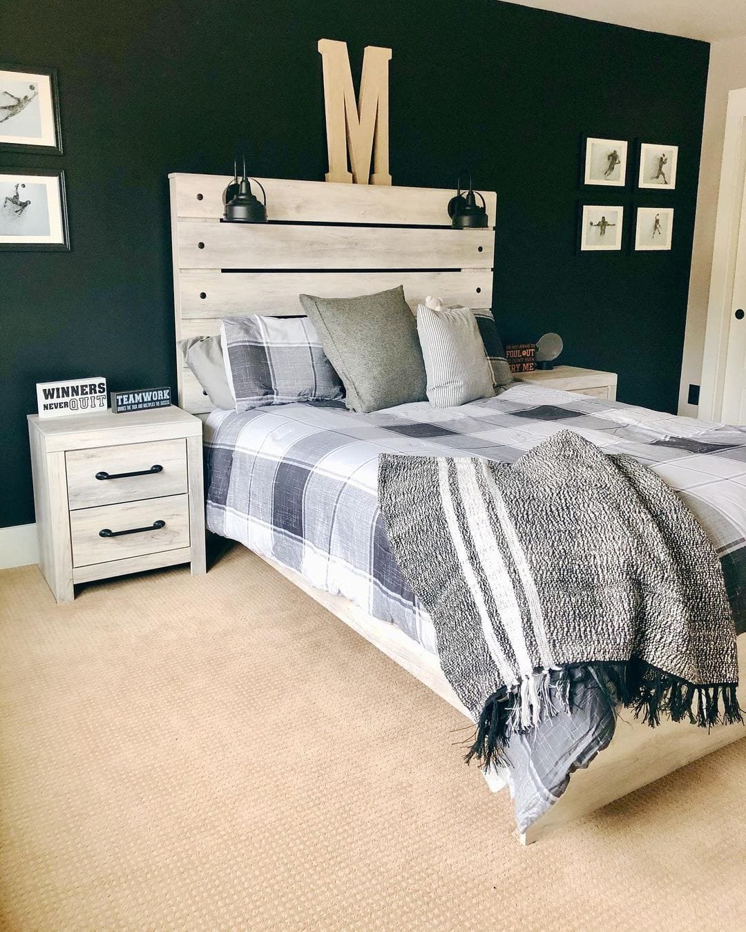 A neatly arranged bedroom with a wooden bed, modern farmhouse bedding, and black walls, decorated with framed pictures and a letter 'M' above the bed.