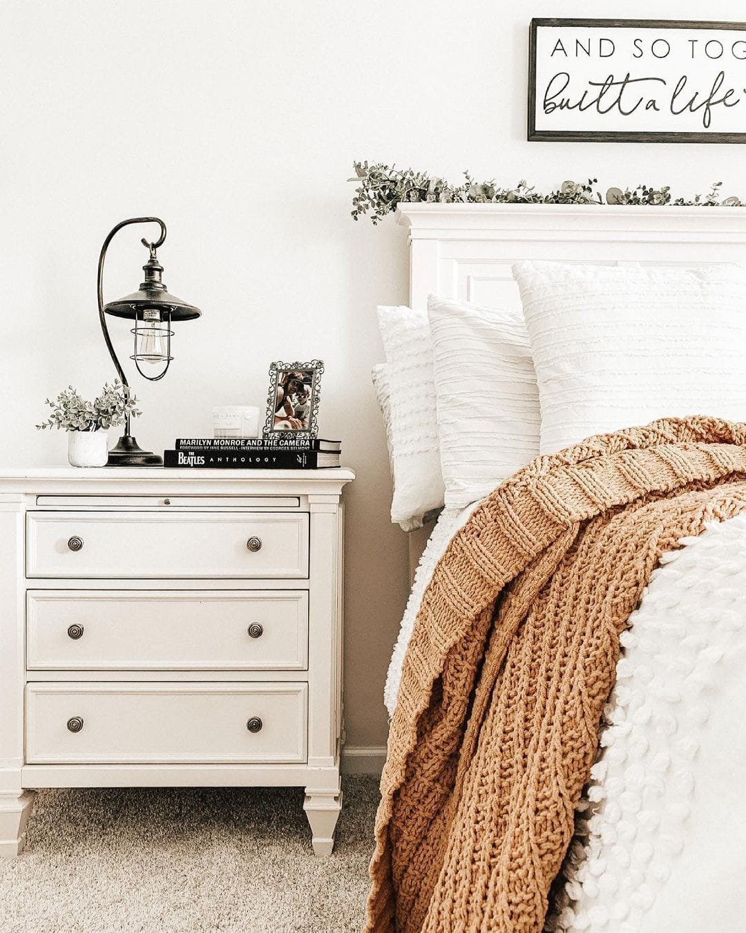 A cozy bedroom corner with a white bed and nightstand, topped with a lamp and books, accented by a modern farmhouse bedding and decorative pillows.