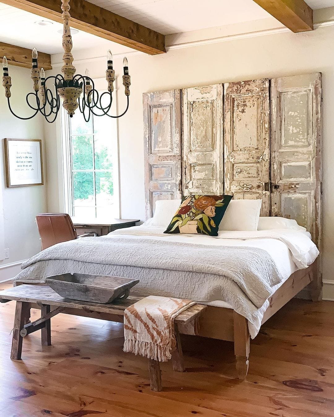 A rustic bedroom featuring a bed with modern farmhouse bedding, a large distressed wooden headboard, a chandelier above, and a hardwood floor.