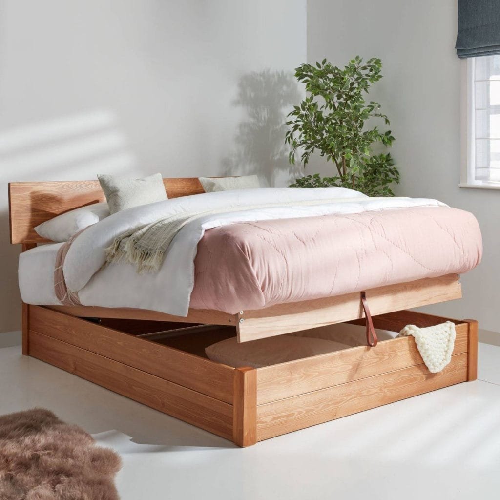 A modern wooden bed frame with a clever pull-out storage drawer, adorned with pink and white bedding, located in a bright room with a plant-shadowed wall.