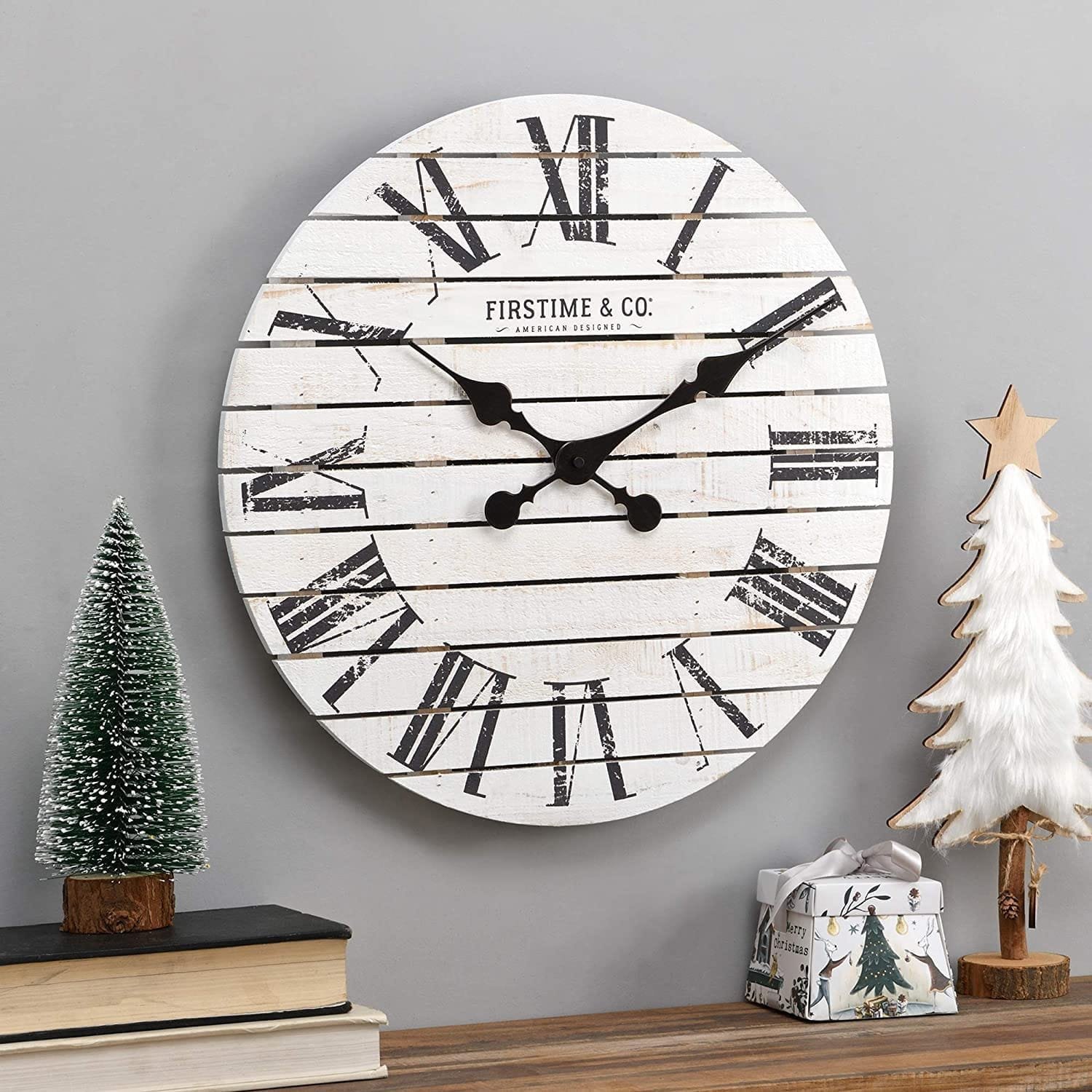 A rustic wall clock with Roman numerals on a white wooden background, flanked by farmhouse decor accents for the living room, including a small green tree and a white tree decoration.
