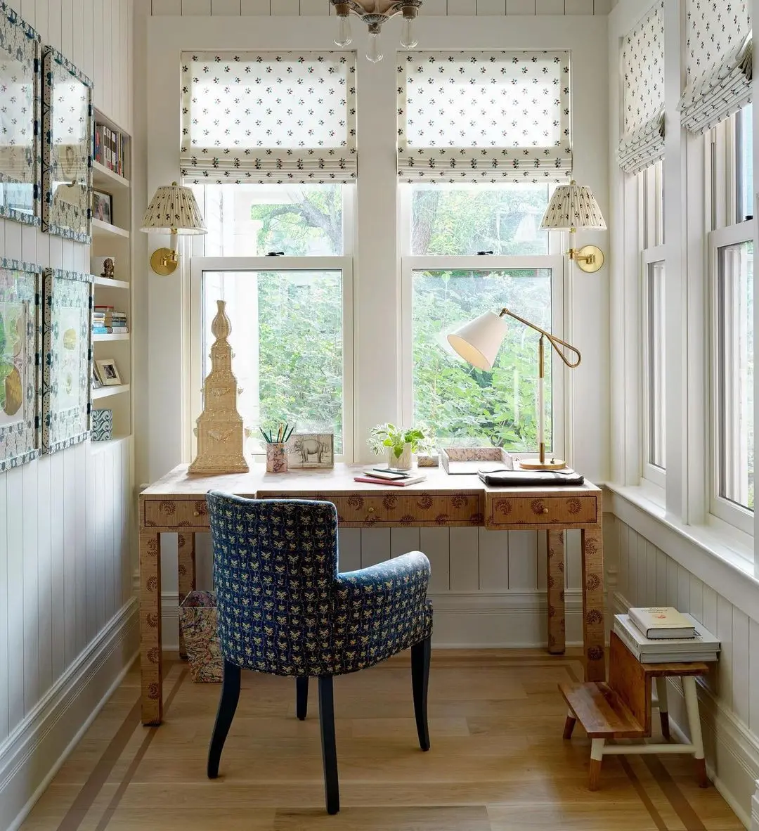 A small, cozy home office with a wooden desk and patterned chair offers maximum efficiency. Large windows with patterned curtains flood the space with natural light, enhancing productivity. Decorative items, plants, and a lamp on the desk create an optimized environment for work.