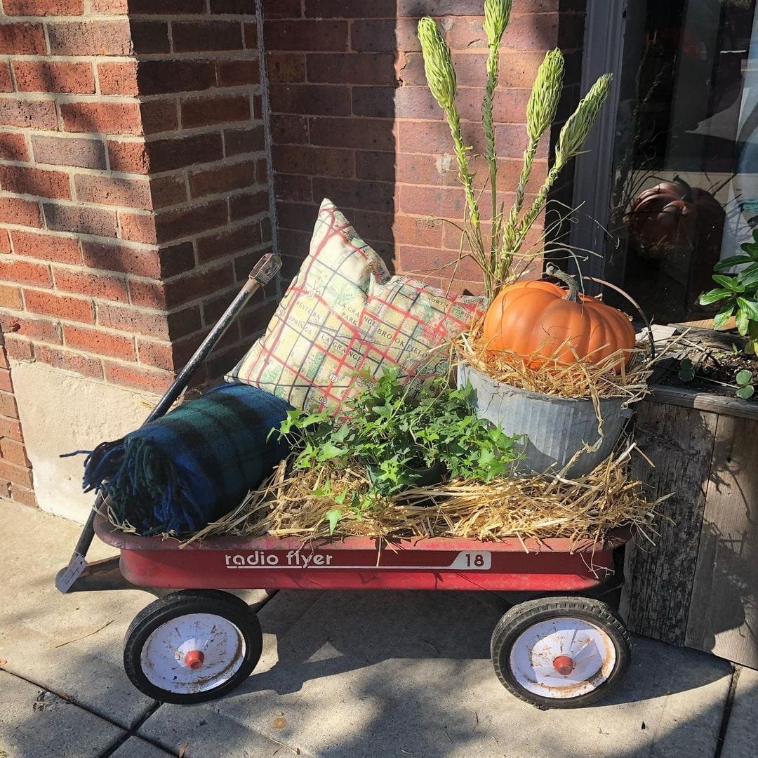 A red Radio Flyer wagon filled with a pumpkin, plants, a plaid pillow, a blanket, and straw creates one of the unique vignettes by a brick wall to decorate an amazing front porch.