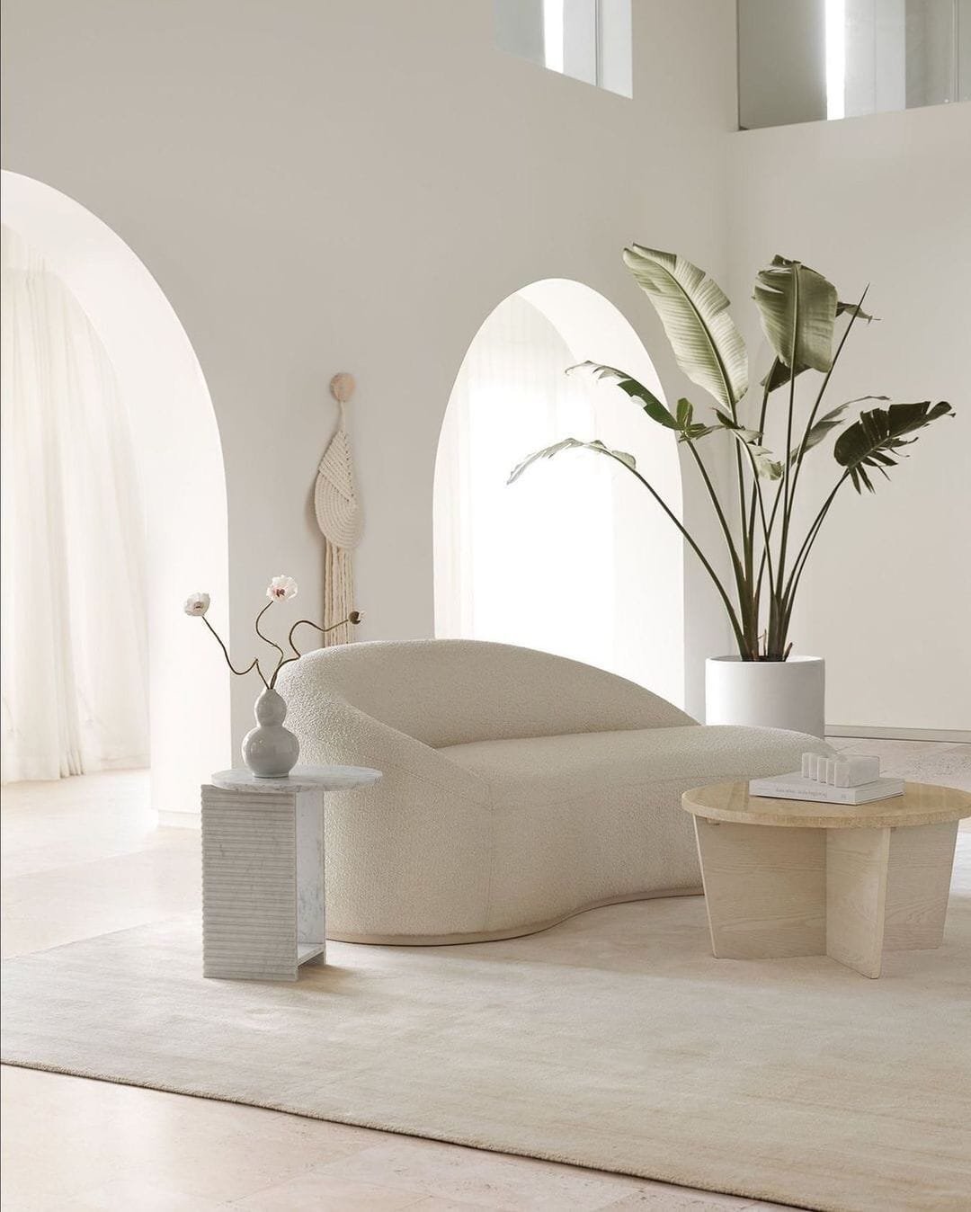A minimalist living room with a plush white sofa, a small side table with decorative items, and a potted tropical plant, set against arched white walls and sheer curtains. Discover our top 10 tips for creating this serene space.