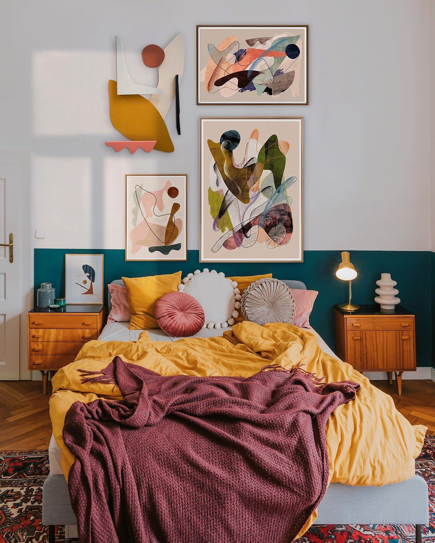 Colorful, artistic bedroom featuring ideas for creating a stunning accent wall with abstract paintings above a bed dressed in yellow and burgundy linens, flanked by lamps on side tables, and a blue half-wall.