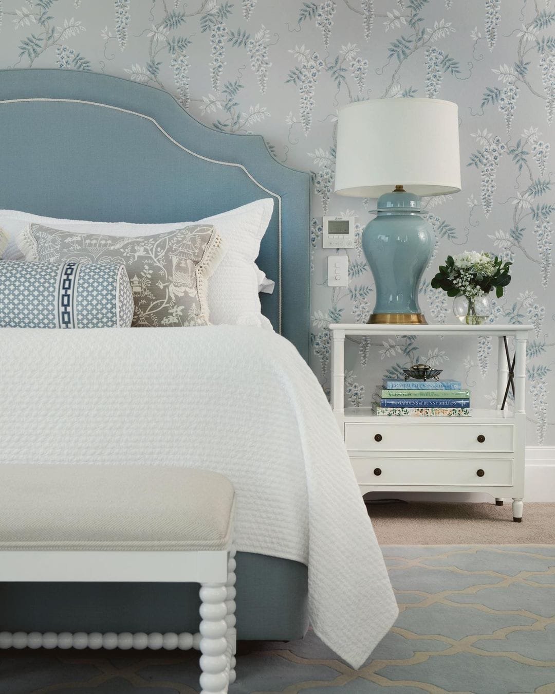Elegant bedroom featuring a blue upholstered bed, stunning accent wall with floral wallpaper, a white bedside table with a lamp, and a decorative vase with flowers.