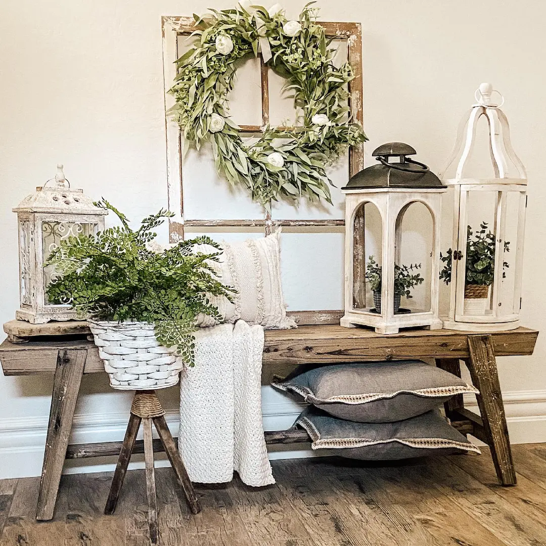 A rustic wooden bench with pillows, a throw blanket, potted plants, lanterns, and a wreath on a repurposed window frame—discover how to style your entryway with a farmhouse bench for the perfect welcoming touch.