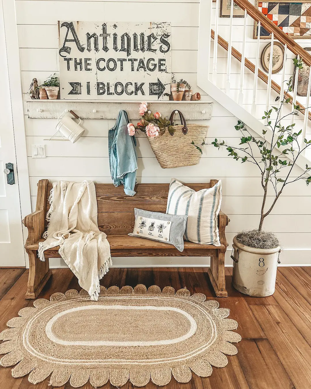 A rustic entryway with a wooden farmhouse bench, pillows, blanket, woven rug, potted plant, and a shelf displaying a vintage “Antiques” sign—perfect inspiration if you’re wondering how to style your entryway with a farmhouse bench.