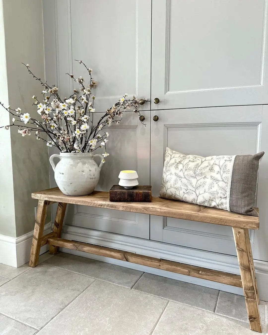 A wooden bench with a floral pillow, two stacked books, and a ceramic pitcher holding white blossoms shows how to style your entryway with a farmhouse bench in front of gray paneled cabinets.