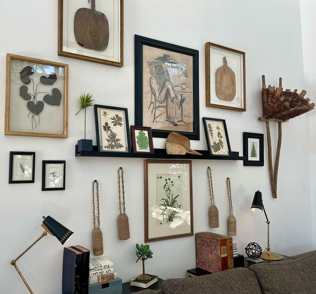A well-decorated living room wall with a variety of vintage frames and artwork, shelves with plants and books, and ethnic decor items.