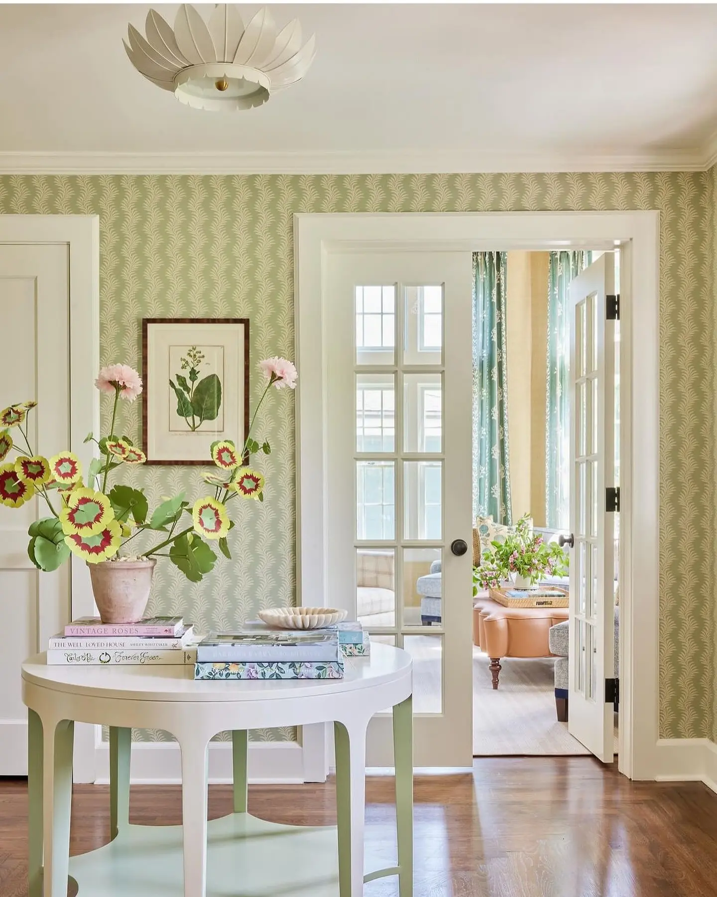 A round white table with books, a bowl, and potted flowers stands in a bright hallway with patterned wallpaper and glass doors, perfectly capturing summer colors: how to incorporate bright hues into your home decor.