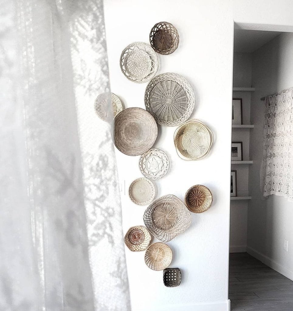 Decorative Bohemian wall baskets of varying sizes and patterns mounted on a white wall in a bright room, with sheer curtains partially visible.