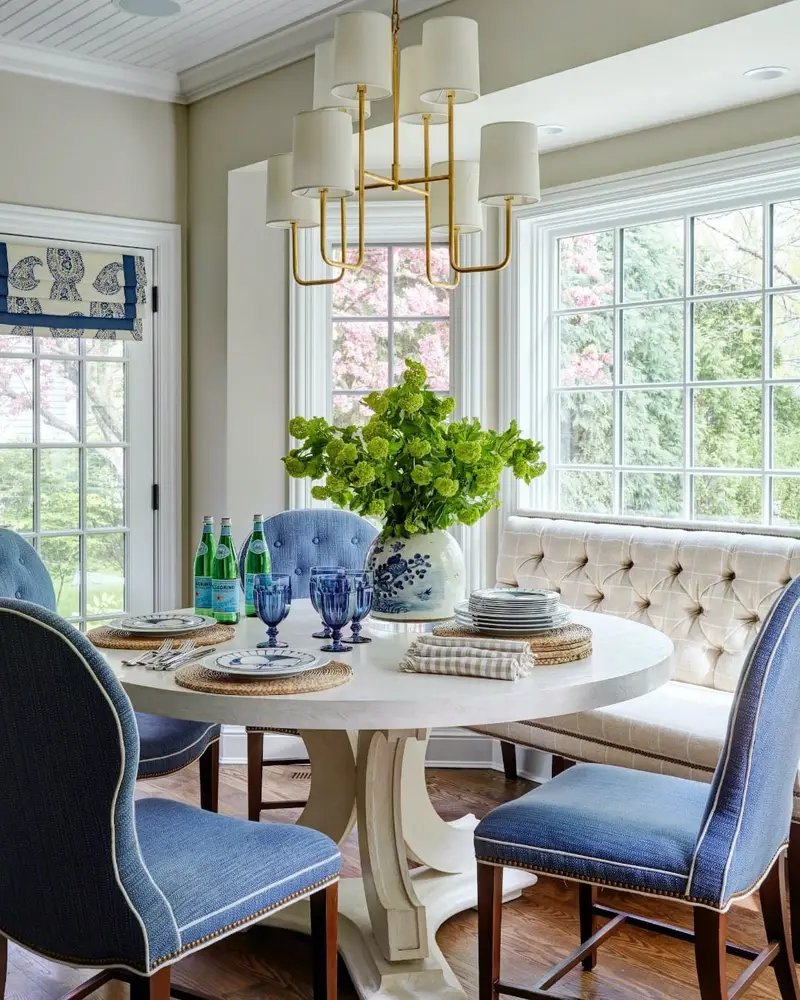 Elegant dining room with a round white table, blue upholstered banquettes, a beige tufted sofa, and a large gold chandelier, surrounded by windows showing green foliage.