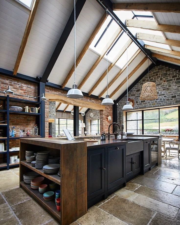 Modern kitchen interior featuring a dark wood island with built-in shelves, gray cabinets, industrial chic exposed brick walls, and high ceilings with skylights.