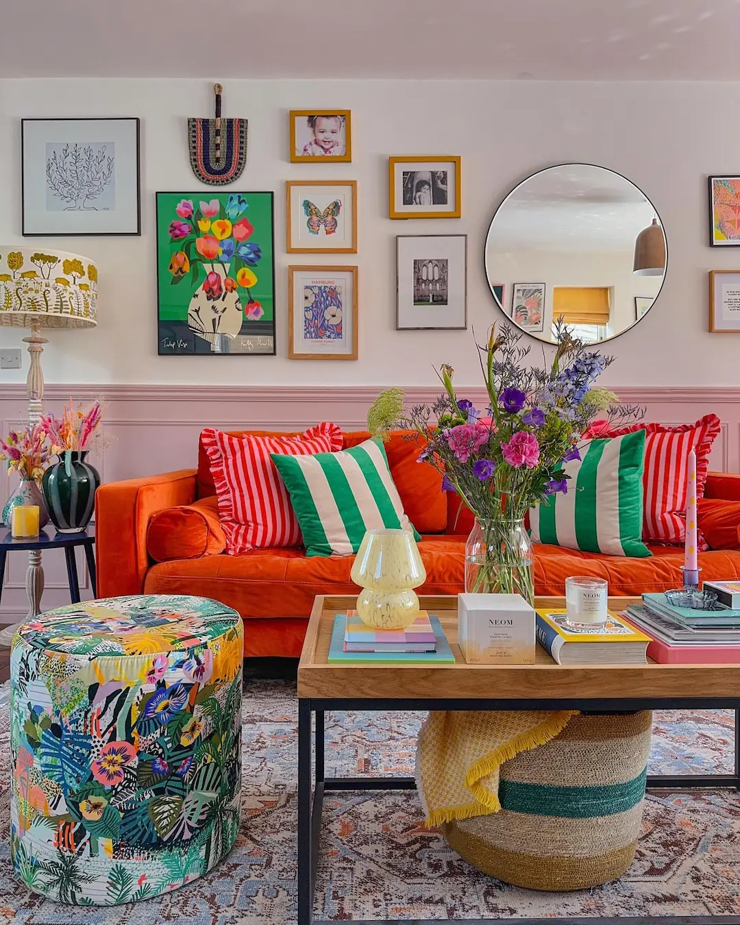 A colorful living room with maximalist decor: an orange sofa, green and red pillows, a floral ottoman, a round mirror, and a gallery wall above the couch show how to balance eclectic style without feeling overwhelming.