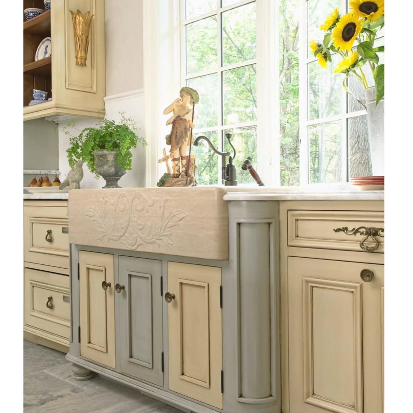 A kitchen with cream-colored cabinetry, a stone farmhouse sink, a potted plant, a decorative figure, and sunflowers in a vase by the window showcases top essential design elements for a French Country Style kitchen.