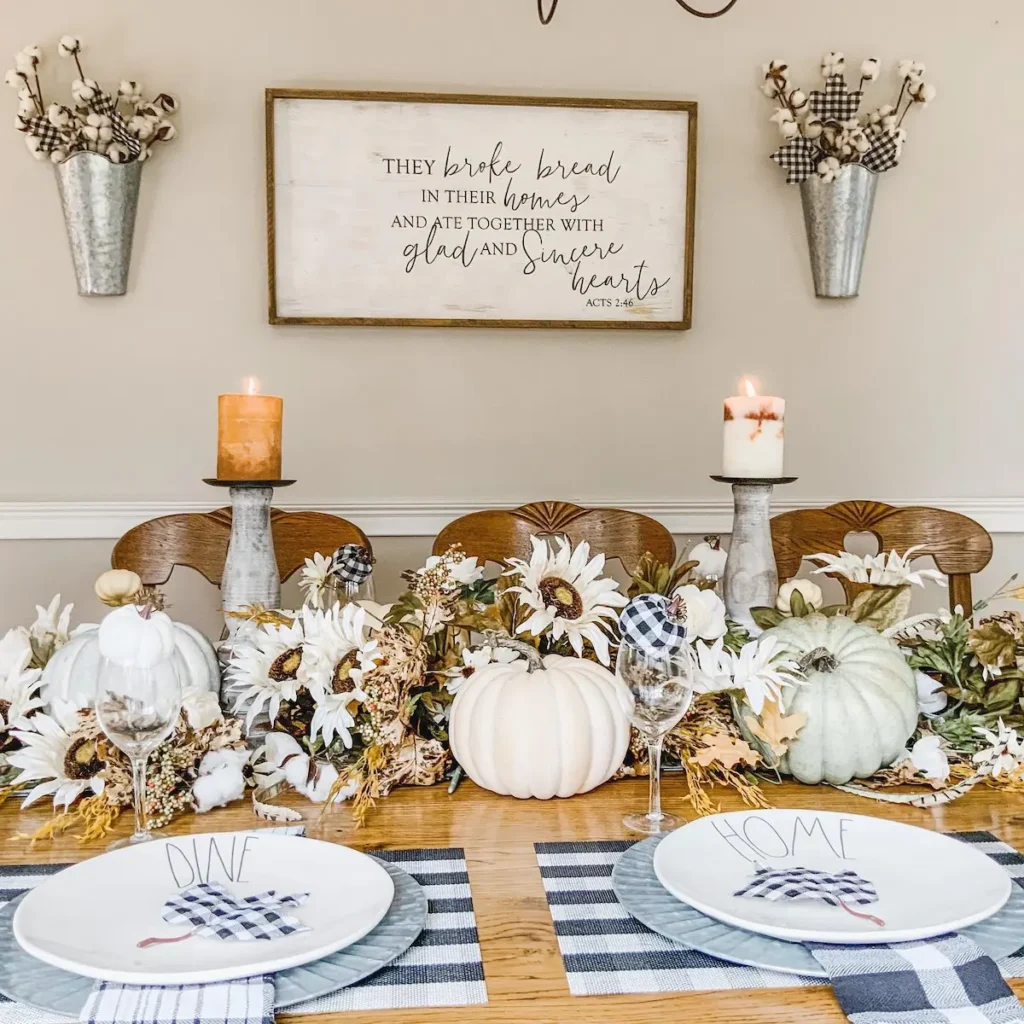 A dining table is decorated with white pumpkins, candles, and floral arrangements. To embrace buffalo plaid in fall decor, add plaid napkins or a runner for a cozy seasonal touch. Two place settings with "DINE" and "HOME" plates sit in front.