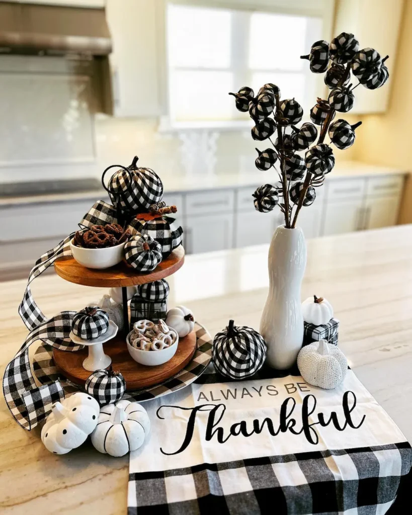 Black and white plaid pumpkins, a vase with matching plaid baubles, and a tiered tray decorated for fall show how to embrace buffalo plaid in fall decor, arranged on a kitchen counter next to a towel that says "Always be Thankful.