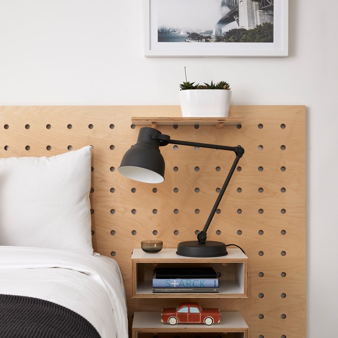 A minimalist bedroom features a pegboard headboard, adding organization made stylish, complemented by a black adjustable lamp, a shelf with a potted plant, a small framed picture, and a wooden box with books and a toy car underneath.