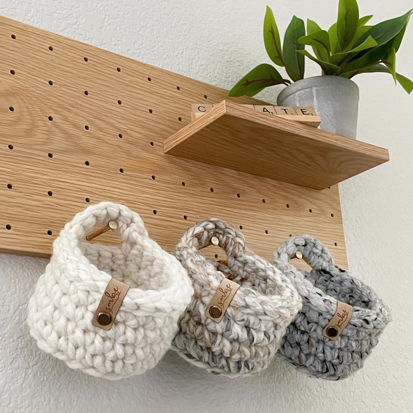 A wooden pegboard shelf holding a potted plant and three small handmade crochet baskets hanging from pegs, each in different shades of cream, multicolor, and gray. Organization made stylish: how to decorate with a wall pegboard.