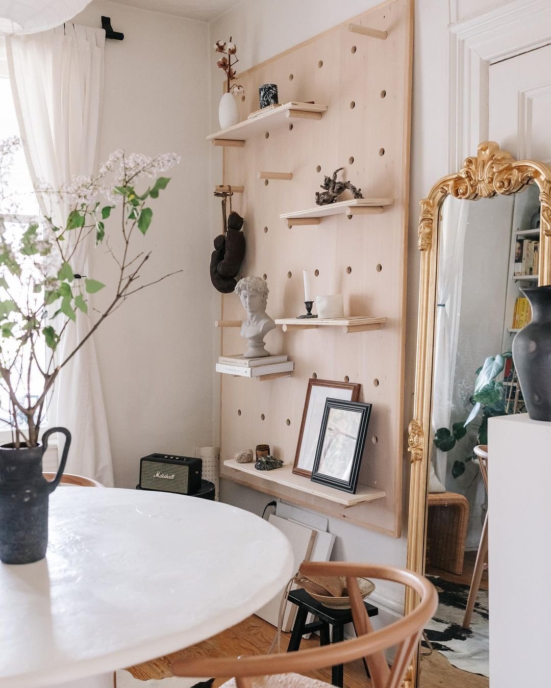 A room features a pegboard shelf with decor items, showcasing organization made stylish. There's a tall ornate mirror with a gold frame, a round white table with a vase of flowers, and a wooden chair. Light streams in from a window with white curtains, creating an inviting atmosphere.