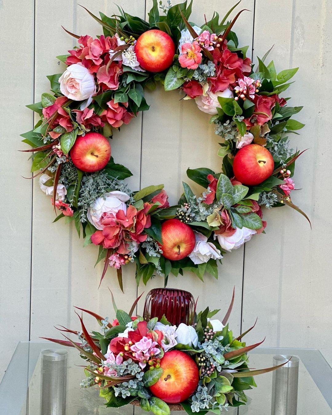 A floral wreath with red apples, pink and white flowers, and greenery hangs on a wooden wall above a matching flower arrangement on a glass surface. Discover how to make your own fall wreaths with natural materials and scents to bring warmth to your home decor.