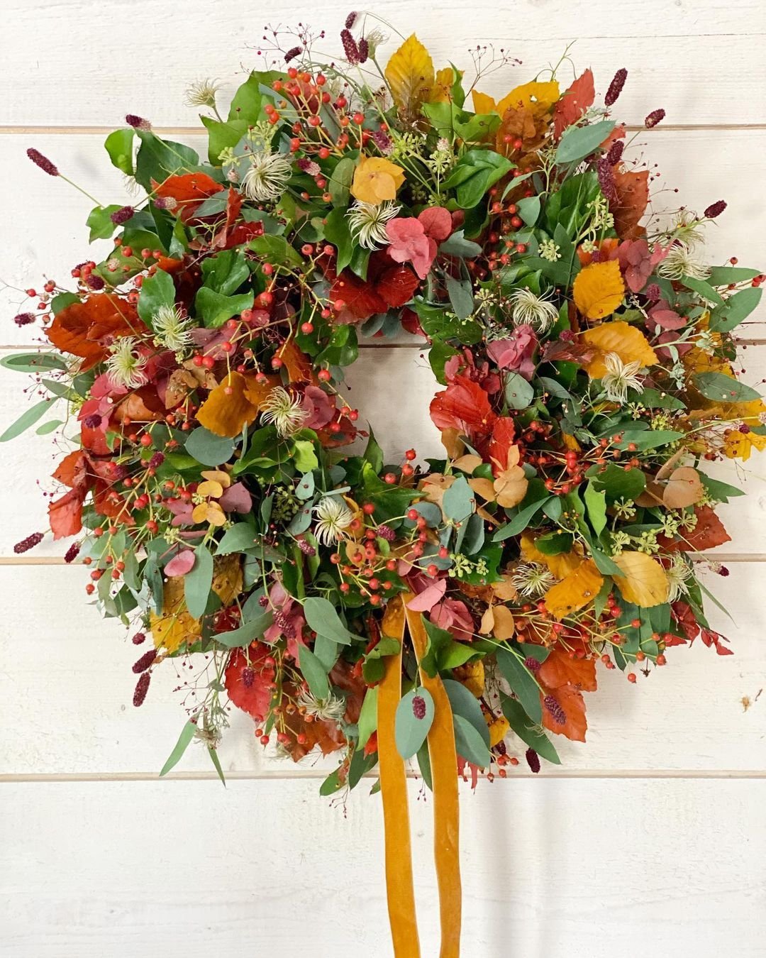 A wreath made of autumn leaves, flowers, and berries, adorned with green foliage and a yellow ribbon, hanging on a wall. Learn how to make your own fall wreaths with natural materials and scents to add a personal touch.