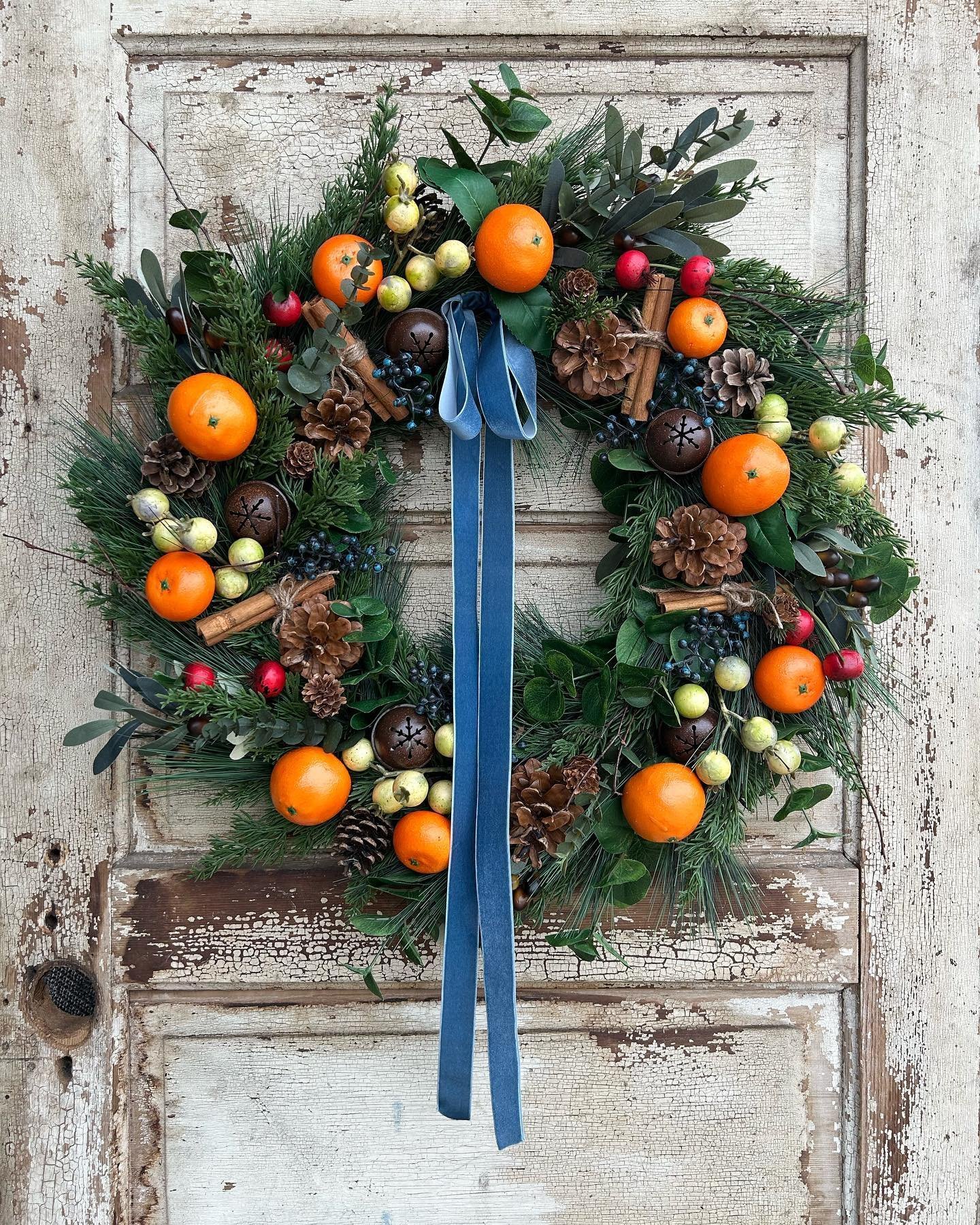 A festive wreath decorated with oranges, pinecones, berries, and green foliage, adorned with a blue ribbon, hangs on a rustic wooden door. Discover how to make your own fall wreaths with natural materials and scents to bring this charming touch to your home.