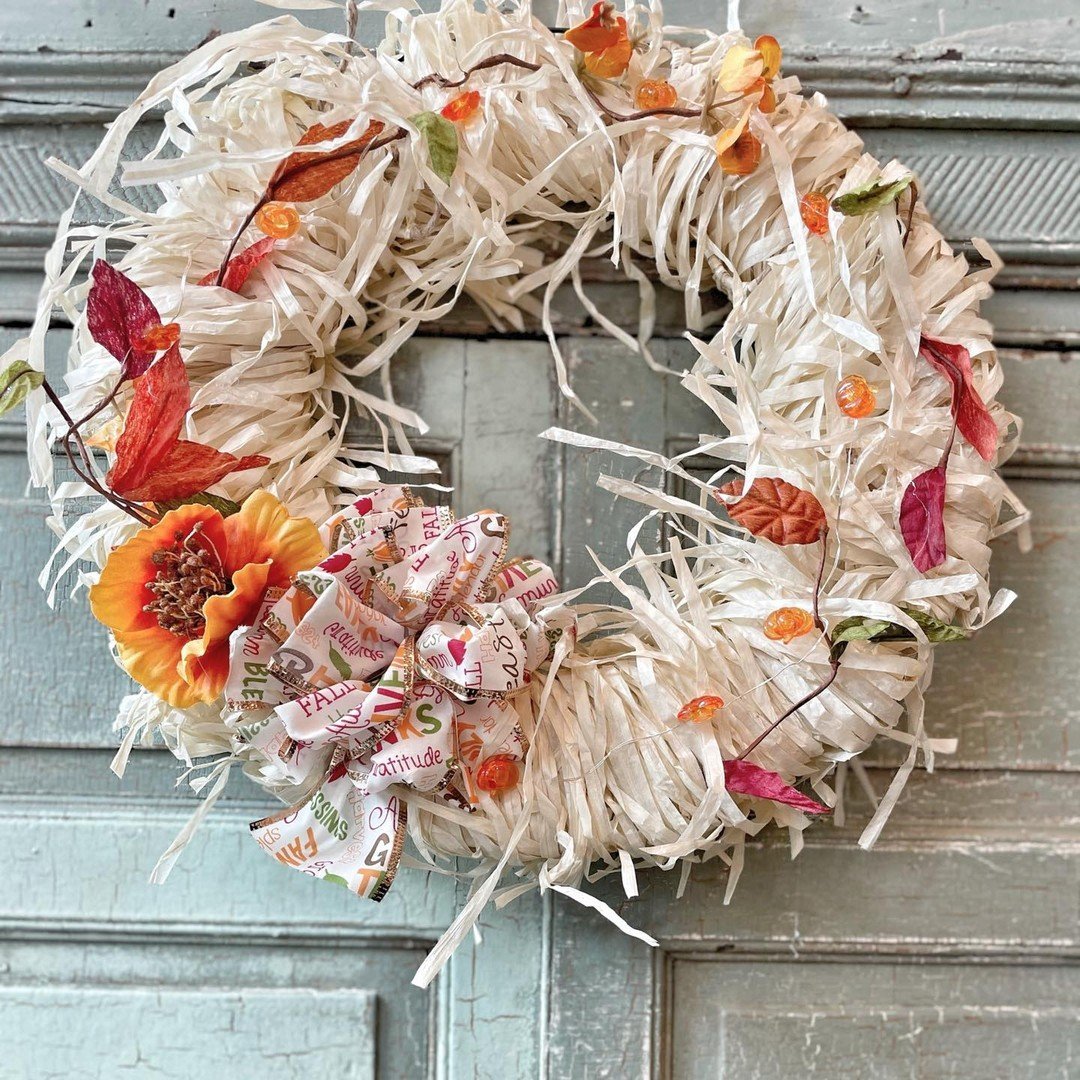 A wreath made of white shredded material, adorned with autumn leaves, a large orange flower, small orange decorations, and a ribbon with words like "fall" and "gratitude," hangs on a rustic wooden door. Learn *how to make your own fall wreaths with natural materials and scents* for an extra touch of seasonal charm.