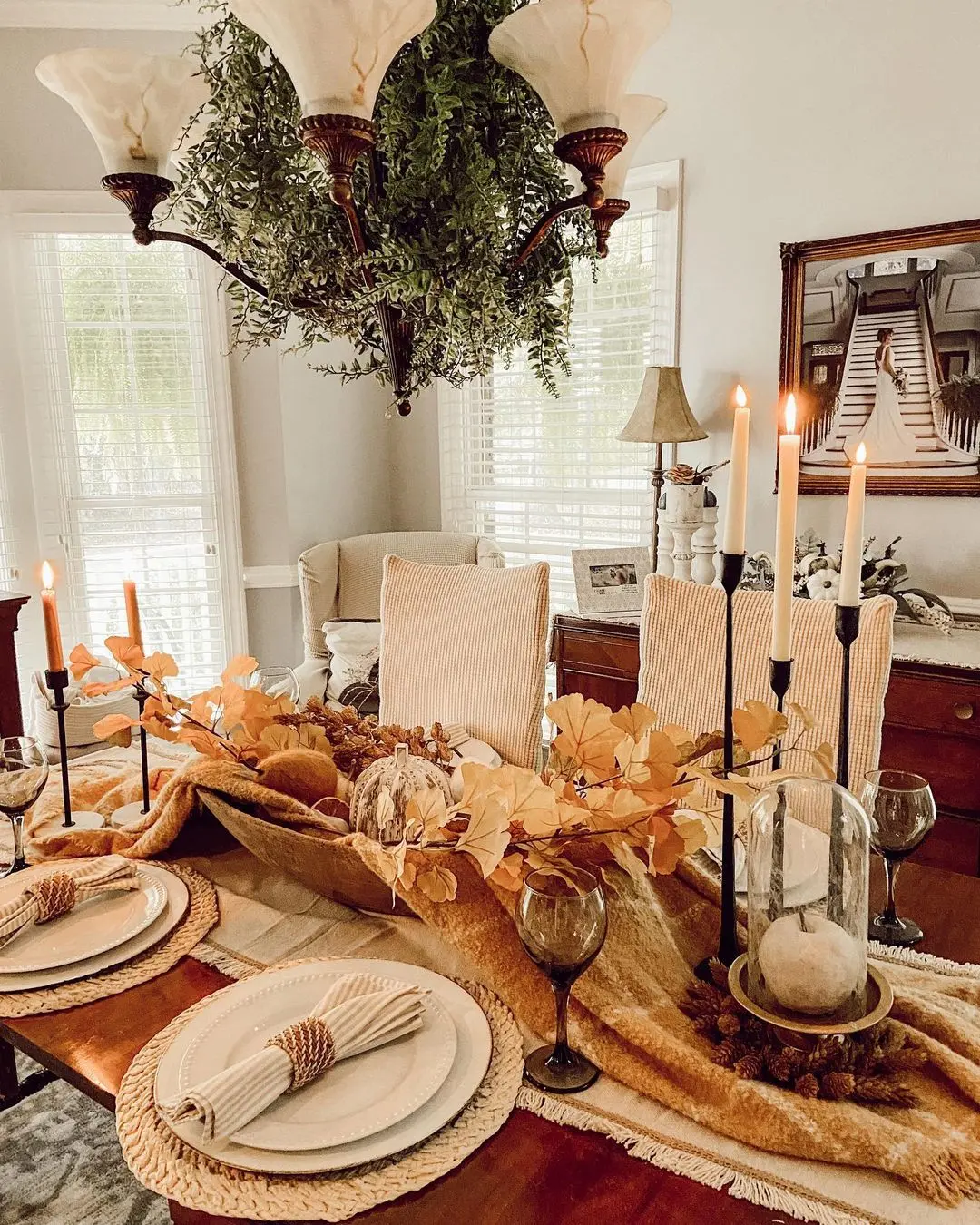 A dining table set for four with autumn-themed decor, featuring candles, gold leaves, woven placemats, and a chandelier adorned with greenery—one of 4 Fall Creative Table Decor Ideas Using Branches and Leaves.