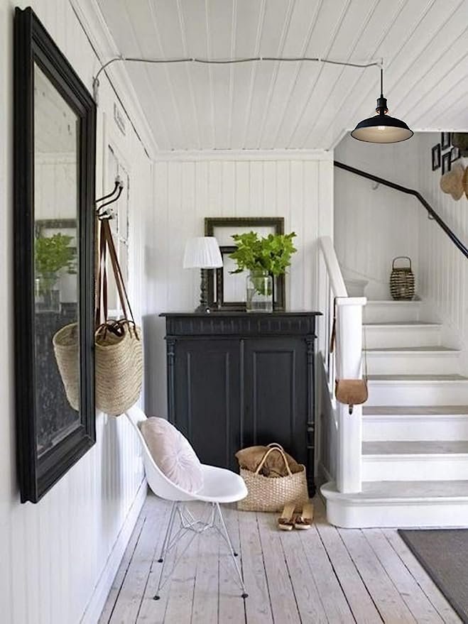 A hallway with white walls and wooden flooring, featuring a black cabinet, a white chair with a cushion, two mirrors, hanging bags, green plants, a staircase, and an industrial barn light. Enchant your home with rustic charm by incorporating this perfect lighting fixture into the space.