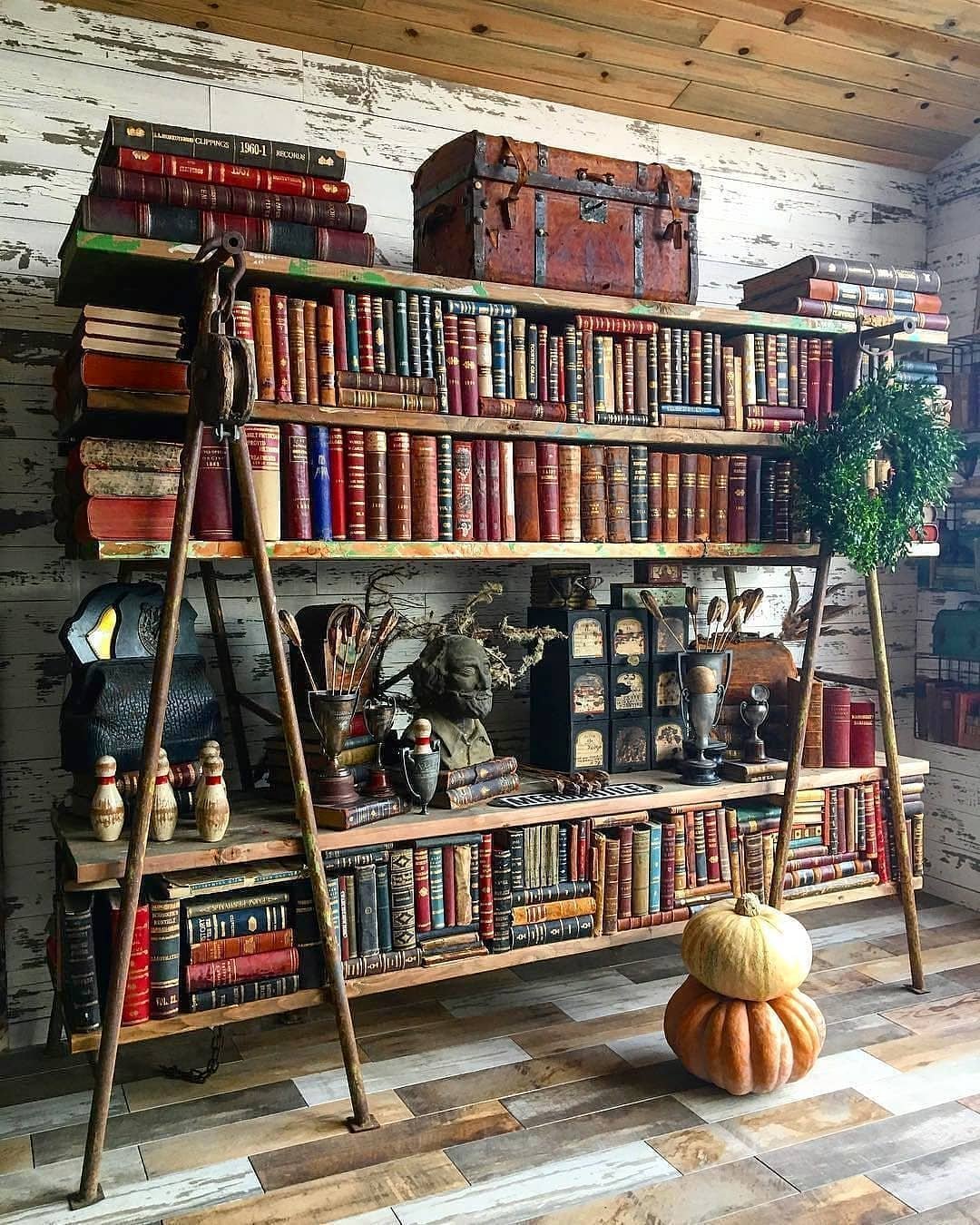 A rustic bookshelf filled with vintage books and various decorative items, including a pumpkin, bowls, a statue, and a green wreath, all against a backdrop of distressed wooden walls. This setting could easily be one of the 10 Inspiring Home Office Library Designs for Every Decor Style.