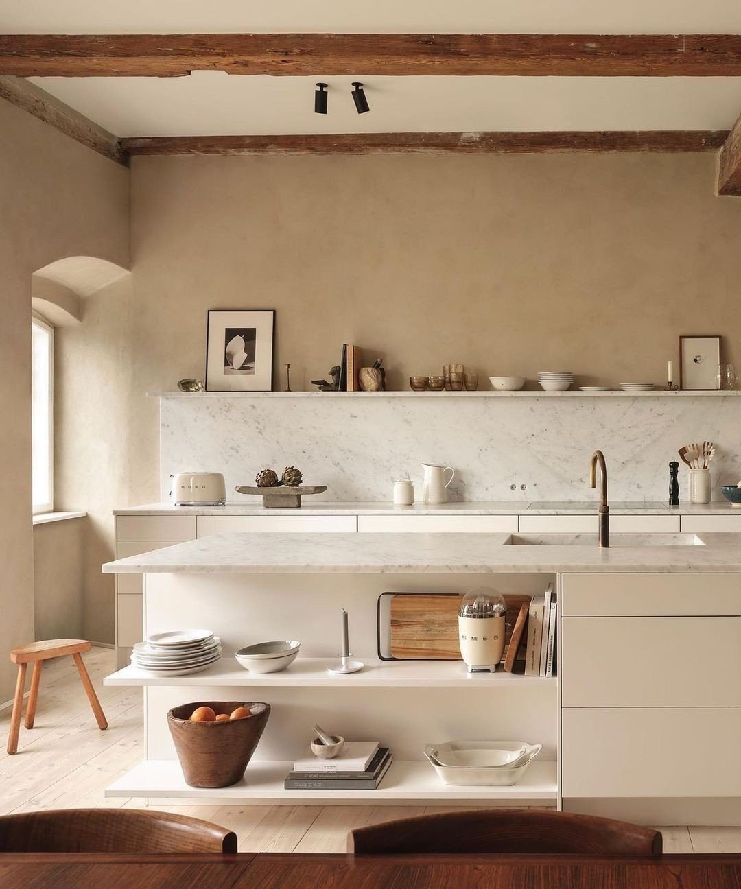A modern kitchen with beige walls, a marble countertop, wooden beams on the ceiling, open shelving with dishware, and minimalistic decor. The Japandi-inspired design creates a functional and beautiful space, complemented by a table with stools in the foreground.