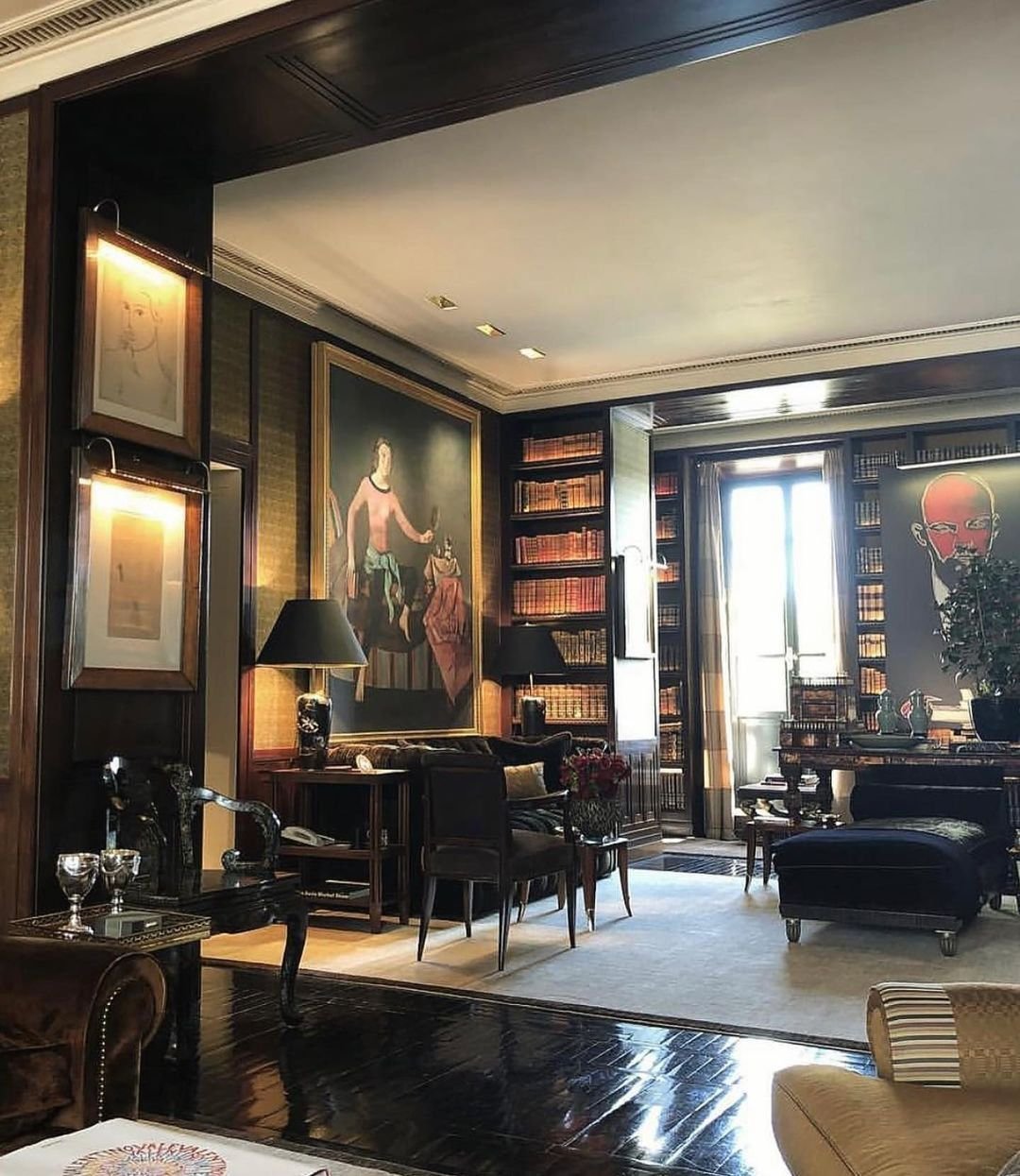 A well-furnished living room with wooden floors, bookshelves, large paintings, and a mix of classic and modern decor. Two lamps illuminate a central seating area with a desk and chairs, blending seamlessly to inspire ideas for 10 Home Office Library Designs for Every Decor Style.