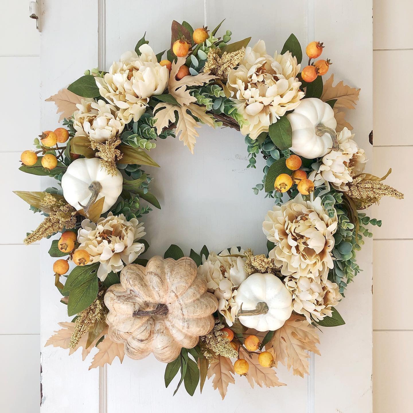 A fall wreath featuring white and beige pumpkins, orange berries, cream-colored flowers, and green foliage hangs on a white door. Get inspired by how to make your own fall wreaths with natural materials and scents for an added personal touch.