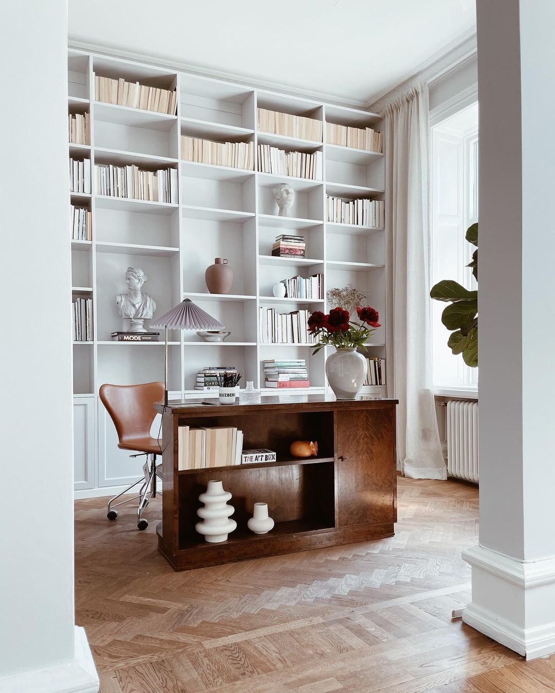 A study room with a wooden desk, modern brown chair, bookshelf filled with books and decorative items, a lamp on the desk, and a large window with light curtains in the background—truly one of the 10 Inspiring Home Office Library Designs for Every Decor Style.