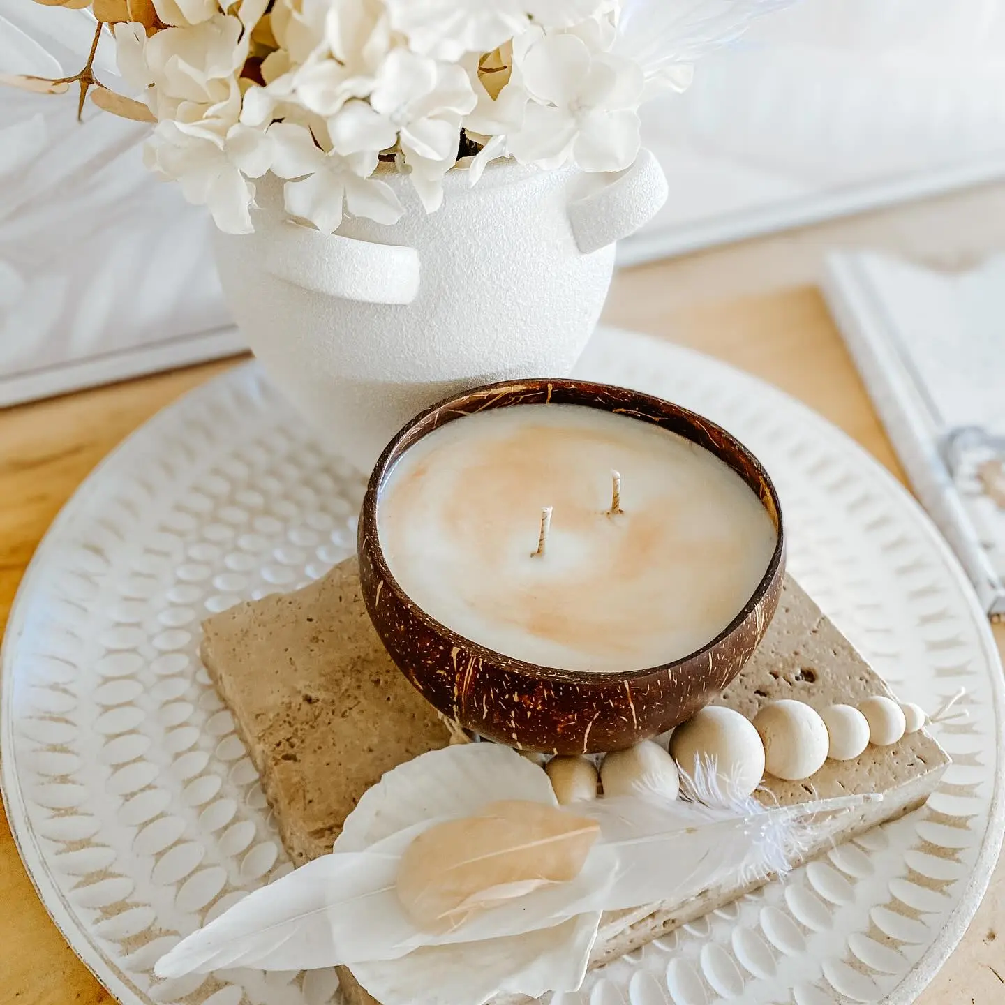 A coconut shell candle with two wicks sits on a stone slab with shells and a feather, next to a white vase of flowers—an inviting setup that shows how to create a calm bedroom with aromatherapy and fall seasonal scents.