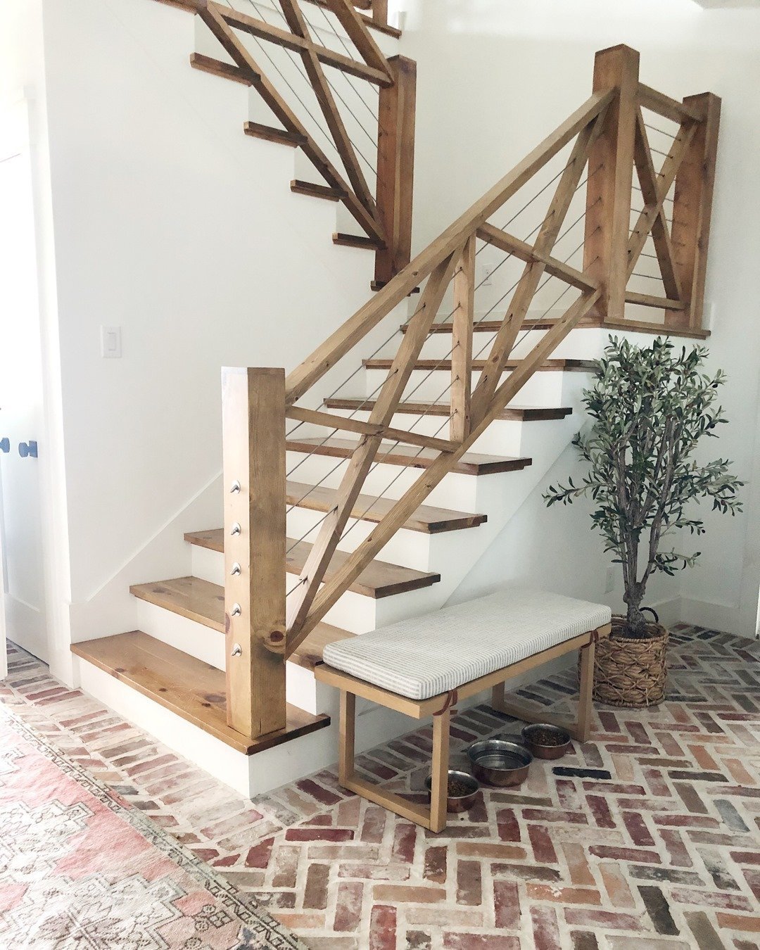 A wooden staircase with an innovative twist features a cushioned bench at the base, adjacent to a small potted tree. The floor has a brick pattern, and there are two pet bowls near the bench.