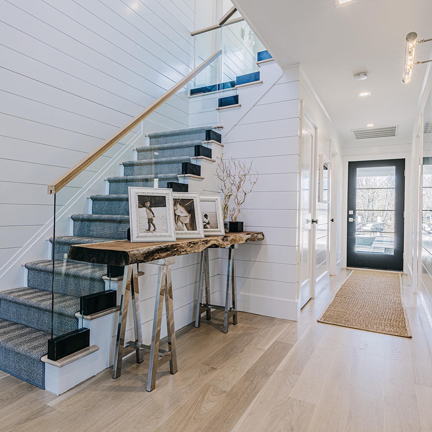 A modern entryway features a wooden table with framed photos and a decorative branch, a staircase with blue carpet showcasing innovative twists on traditional farmhouse railing designs, white shiplap walls, and a black door with glass panels.