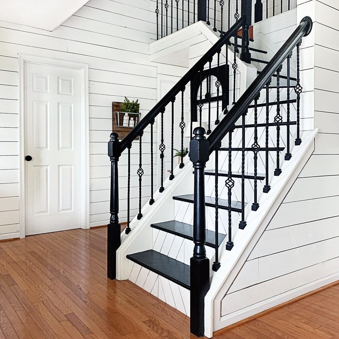 A wooden staircase with black rails and white steps in a house with white shiplap walls and hardwood floors showcases innovative twists in farmhouse stair railing designs.