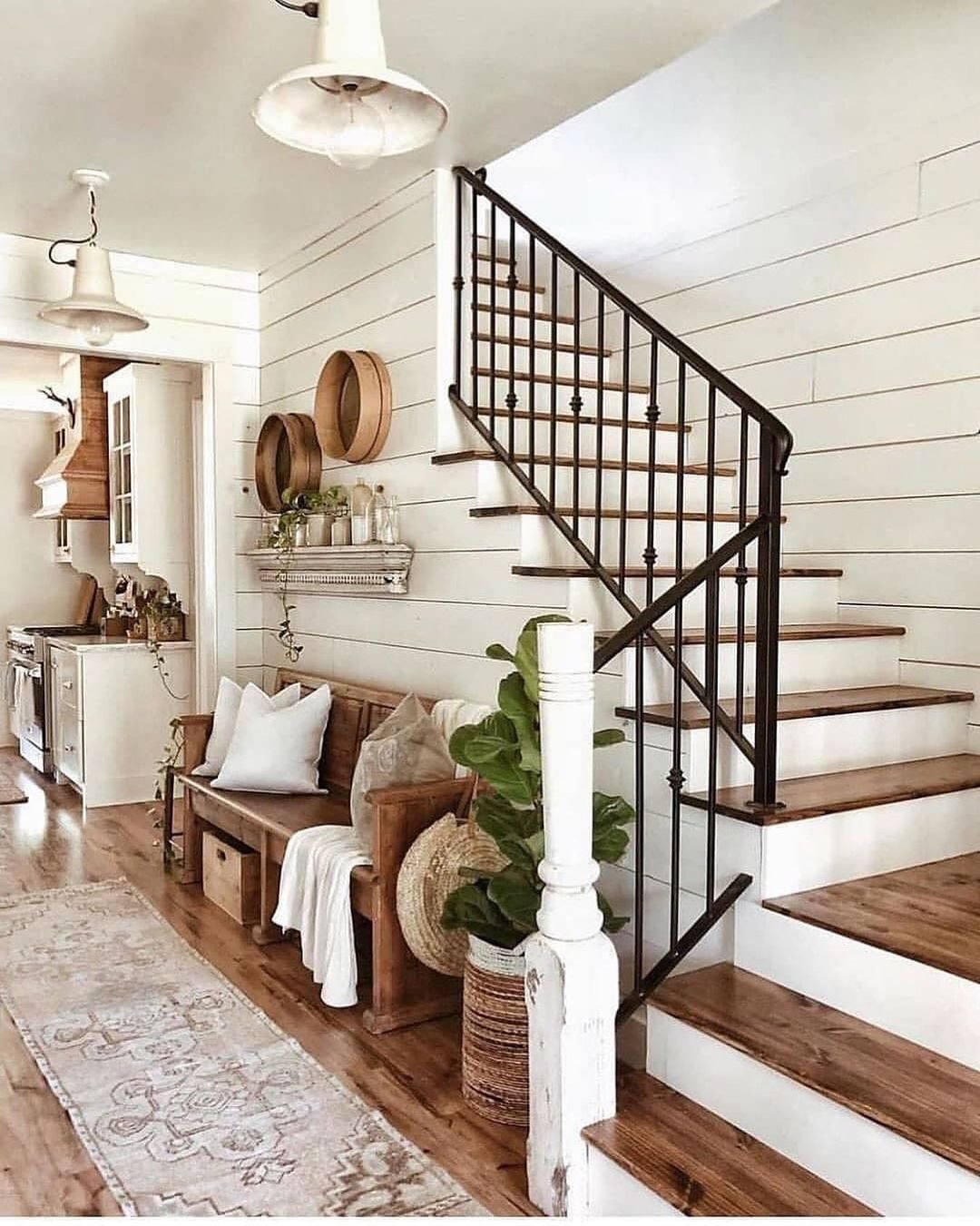 A cozy farmhouse-style entryway with a wooden bench, plants, and wall-mounted baskets beside a staircase featuring black metal railing. Innovative twists like these unique ideas for farmhouse stair railing designs truly enhance the charm. White shiplap walls and wooden floors complete the look.