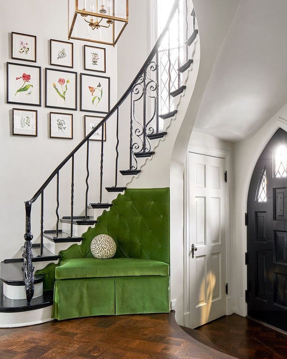 A foyer with a curved staircase showcasing innovative twists in farmhouse stair railing designs, a green cushioned bench under the stairs, a black front door, and framed botanical prints on the wall. The space features dark wood floors and a hanging light fixture.