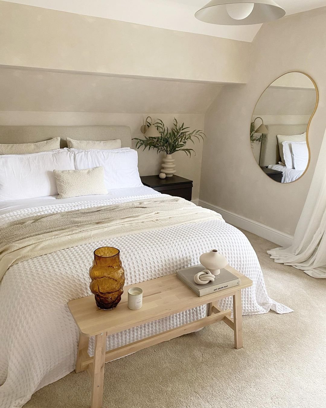 A cozy Japandi bedroom featuring a neatly made bed, plants, a wooden bench at the foot, and an irregular-shaped mirror on the wall. The room uses neutral tones and minimalistic decor to show how to incorporate meaningful artifacts seamlessly.