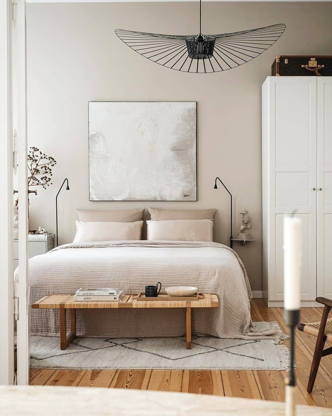 A neatly arranged bedroom featuring a double bed with beige bedding, a wooden bench at the foot, a white wardrobe, abstract wall art above the bed as if following tips on how to incorporate meaningful artifacts in your Japandi bedroom, and a modern black light fixture.