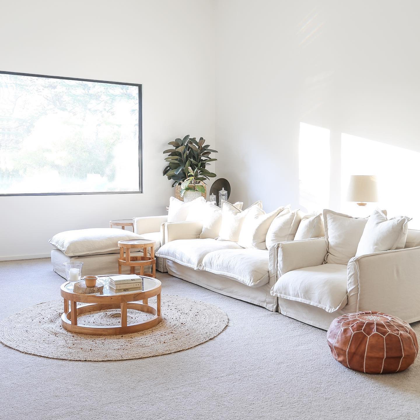 A sunlit living room with a large white slipcover sectional sofa, a circular woven rug, a round wooden coffee table, and a large window with a view. A plant and side table are in the corner, showcasing how slipcover sofas enhance your living room's charm.