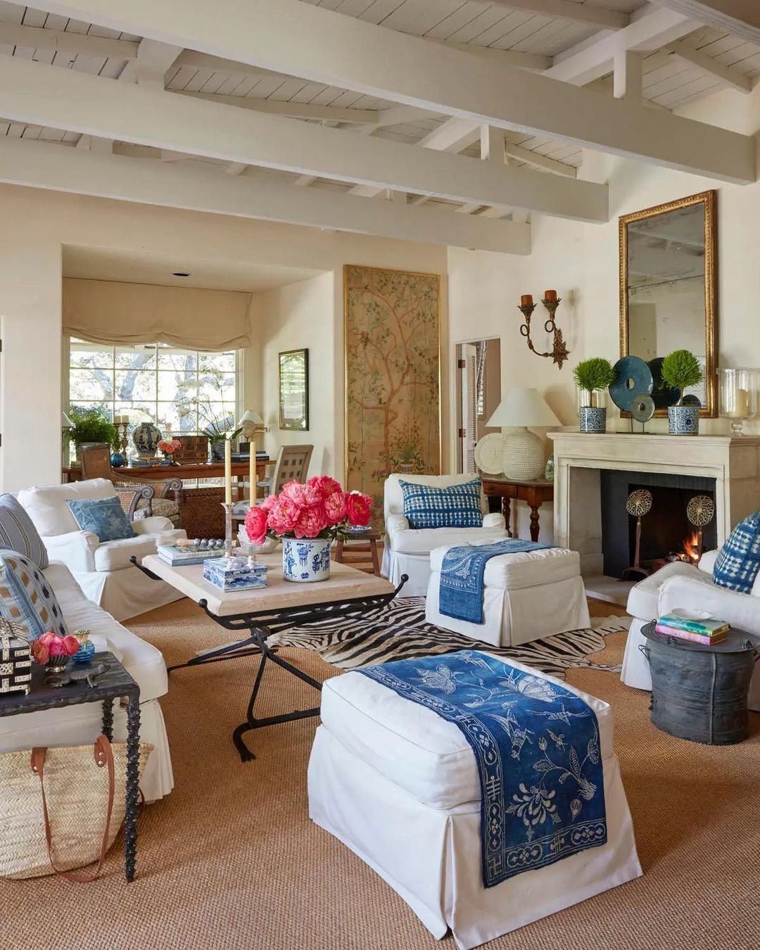 A cozy living room features white slipcover sofas with blue accents, a fireplace, floral arrangements on tables, and a large window allowing natural light. Decor includes various art pieces and a large mirror. Discover how slipcover sofas enhance your living room's charm effortlessly.