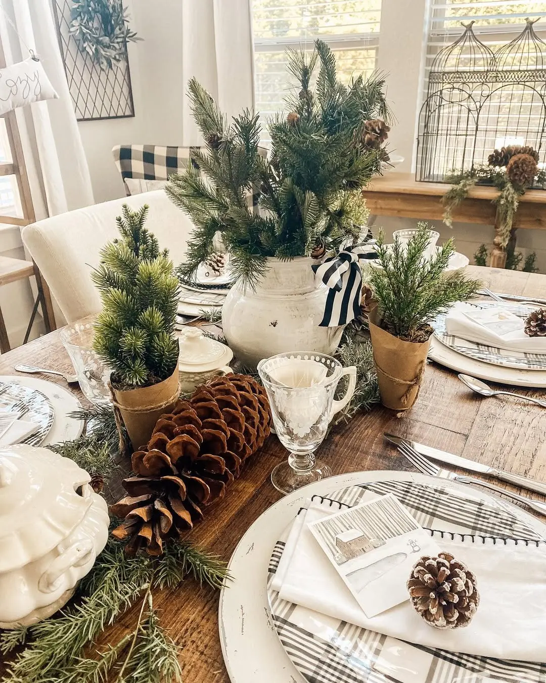 A wooden dining table set with plaid plates, napkins, pinecones, and potted pine trees creates a rustic winter centerpiece—an inviting way to embrace the season and transition your home decor from fall to winter.