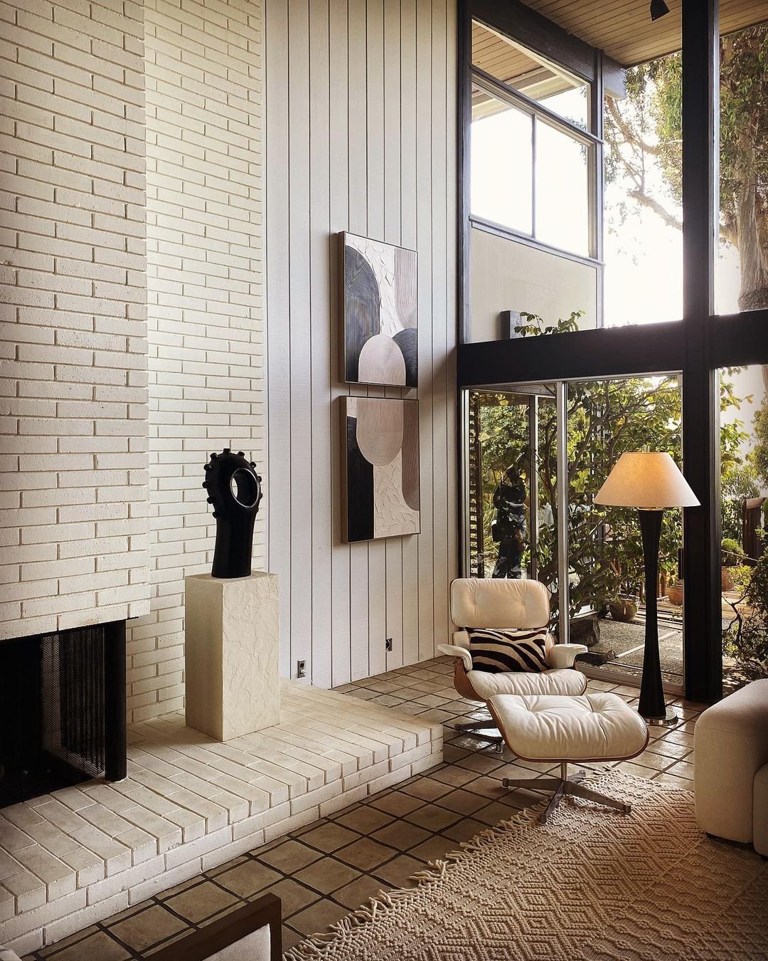 Modern living room with high ceilings, large windows, light brick fireplace, white lounge chair, abstract wall art, and a lamp by glass doors opening to a garden. Minimalistic décor with natural light captures Mid Century Modern Minimalism at its best: Inside Eichler Homes.