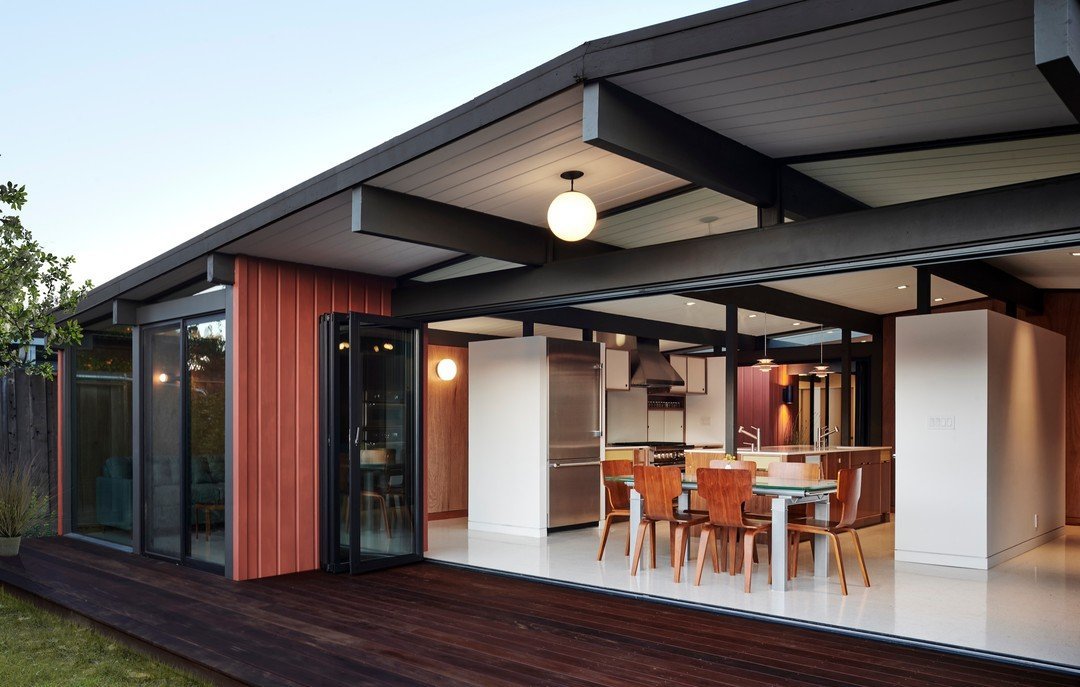 A modern, single-story house with a large open sliding door reveals a contemporary kitchen and dining area. The exterior features wood and glass elements, with a wooden deck extending from the house—truly Mid Century Modern minimalism at its best, reminiscent of Eichler Homes.