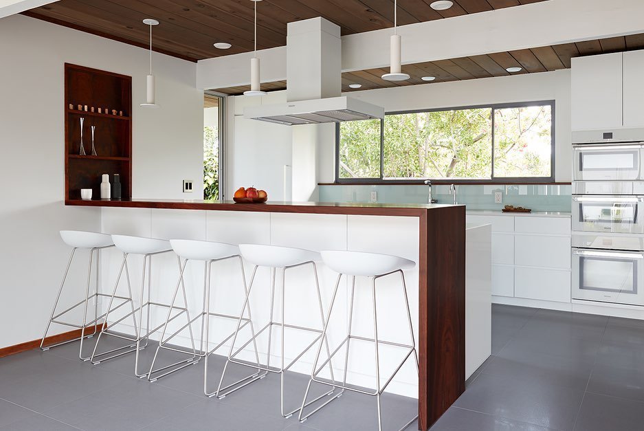 Modern kitchen with white cabinets, a wooden accent island, and four white bar stools. Pendant lights hang from the ceiling. Large window in the background provides natural light. This space echoes why Eichler Homes Interior Design is a mid-century masterpiece with its clean lines and understated elegance.